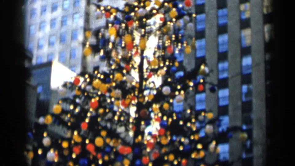 Close, grainy 8mm frame of a New York City Christmas tree with dense ornaments and light points before tall buildings.