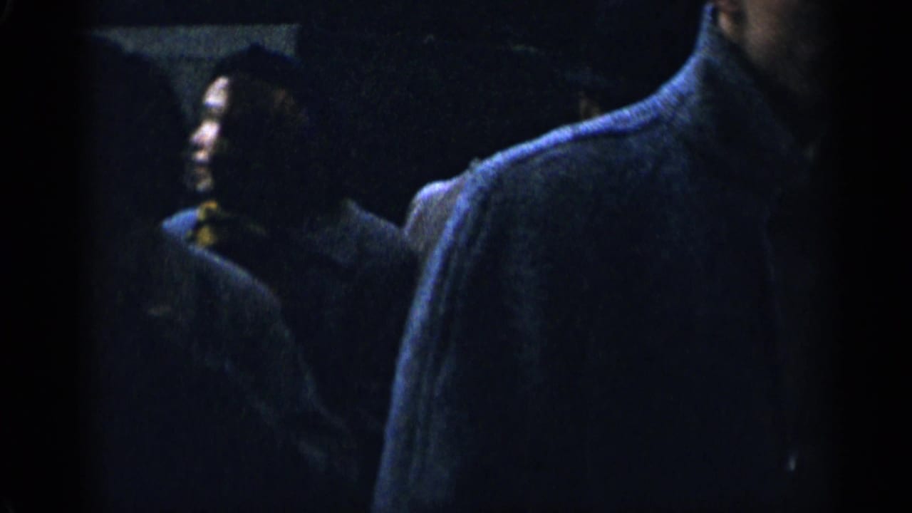 Bundled pedestrians in dark winter coats pass through a dim nighttime street scene.
