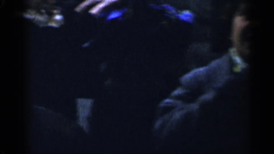 A shadowed 8mm night frame shows a bundled person at right with dim crowd shapes behind them in New York City.