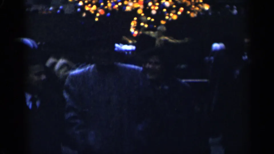 1955 New York City home-movie frame of adults in winter coats moving beneath soft overhead Christmas lights at night.