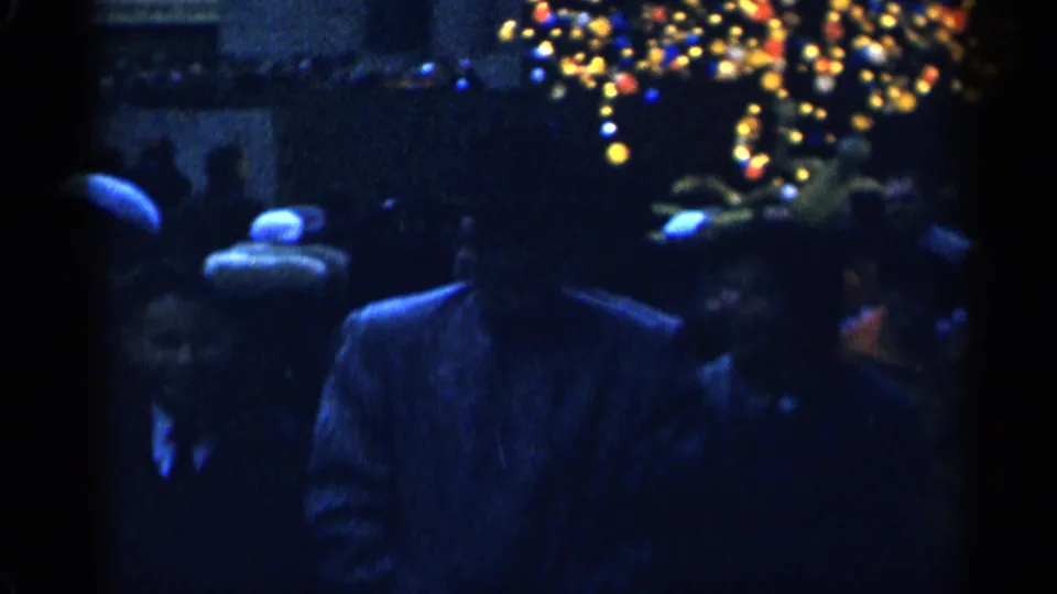 Blue-tinted 8mm frame from New York City showing adults in winter hats and coats beneath glowing Christmas decorations.