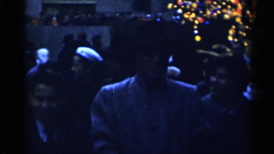 Handheld 1955 New York City holiday frame showing pedestrians clustered under colorful lights in a dark street setting.