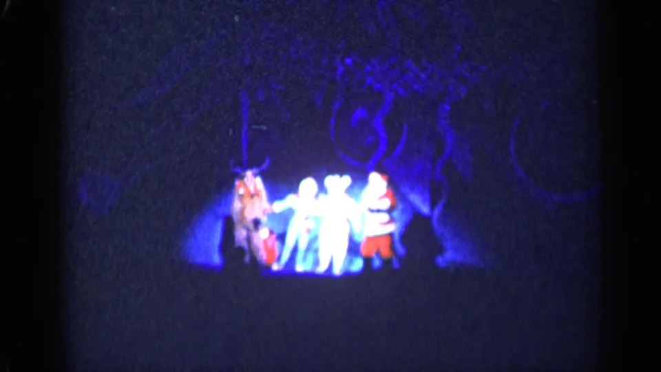 1955 New York City home-movie frame showing a Christmas stage group under blue light with a faint patterned backdrop.