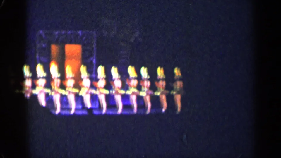 1955 New York City 8mm frame showing a dance troupe lined across a dim stage with glowing yellow and orange costume accents.