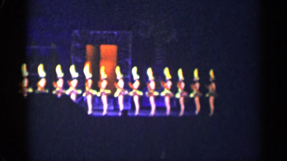 1955 New York City stage-performance frame with dancers in coordinated formation near central orange-lit scenery.