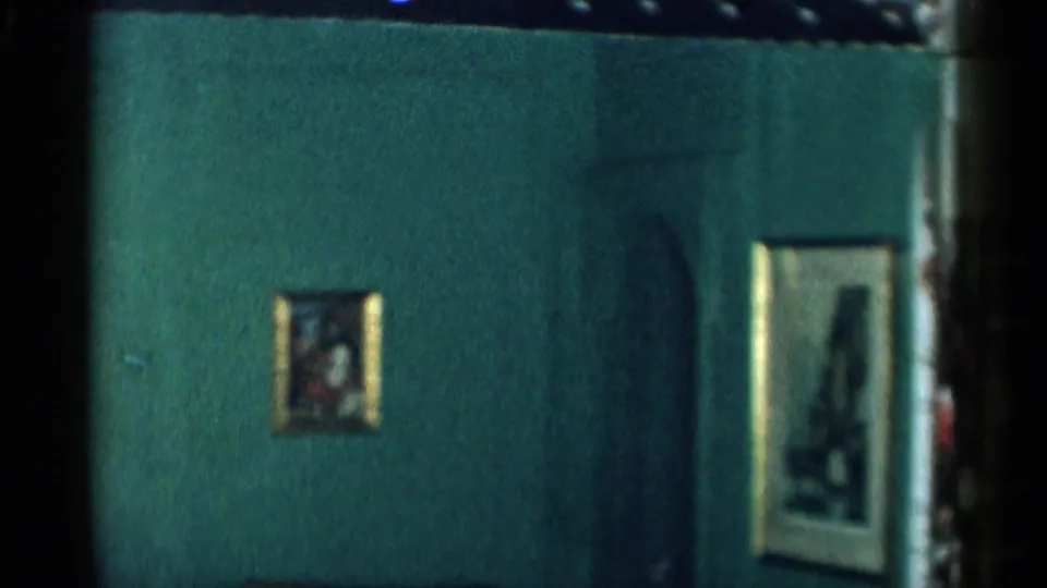 Dark green interior wall with small and larger framed pictures, filmed in a 1955 New York City home movie.