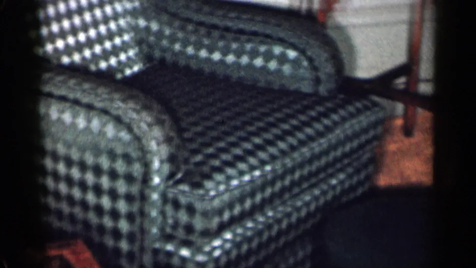 Close 1955 home-movie view of the lower blue diamond-pattern armchair, showing upholstery, wood floor, and background table legs.