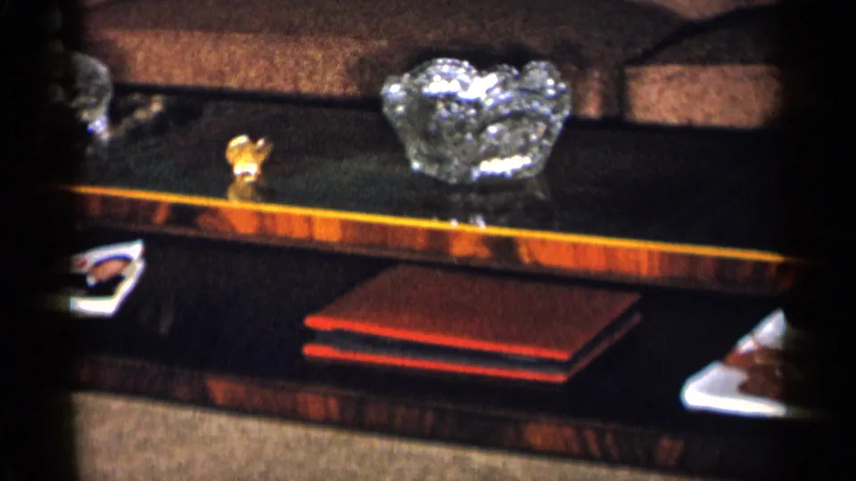 Home-movie frame of a mid-century living-room table with crystal decor, a red book or album, and a couch in the background.