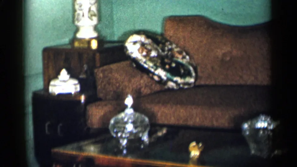 Home-movie view of a private New York City living room with brown couch, patterned pillow, side table, and crystal pieces on the coffee table.