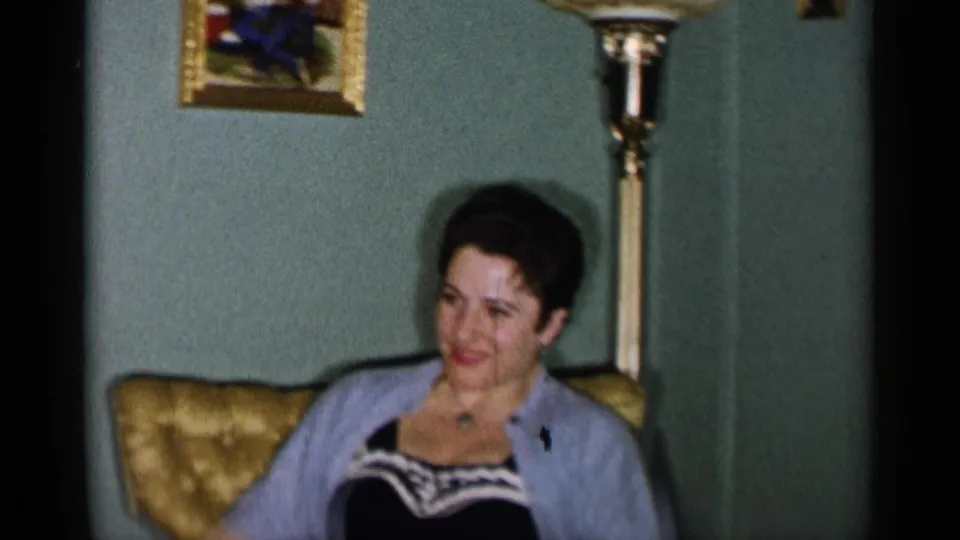 1955 New York City home-movie frame of a woman in a light cardigan seated in a gold tufted chair, with a floor lamp and framed picture behind her.