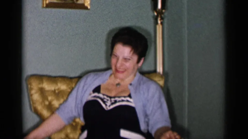 1955 New York City home-movie frame showing a smiling woman seated in a gold tufted armchair, wearing a light cardigan over a dark dress.