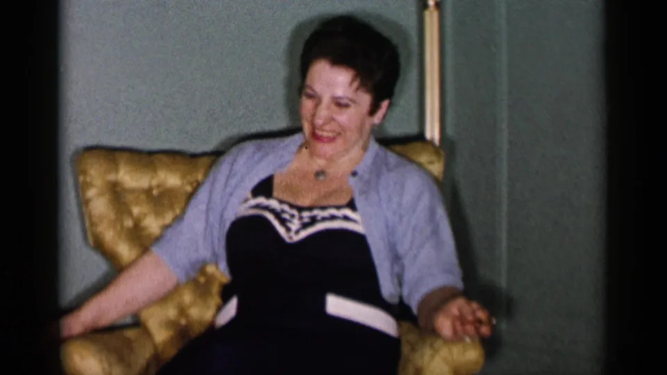 A woman in 1955 New York City home-movie footage laughs in a gold tufted chair, wearing a dark dress and light cardigan.