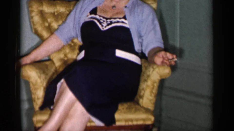 The seated woman shifts in the armchair, with crossed legs and a cigarette visible in one hand inside a private room.