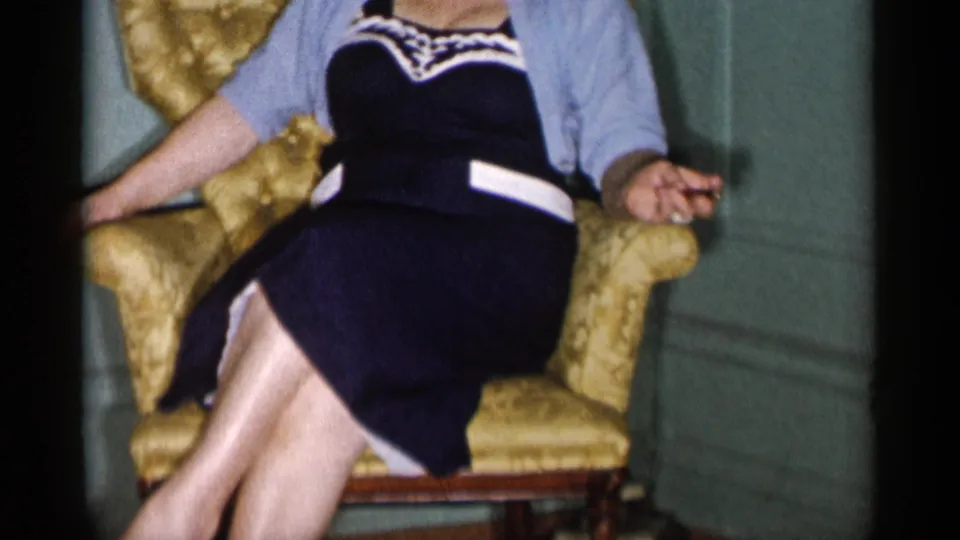 A close, slightly soft home-movie frame shows the woman seated in a gold armchair, her face mostly cropped from view.