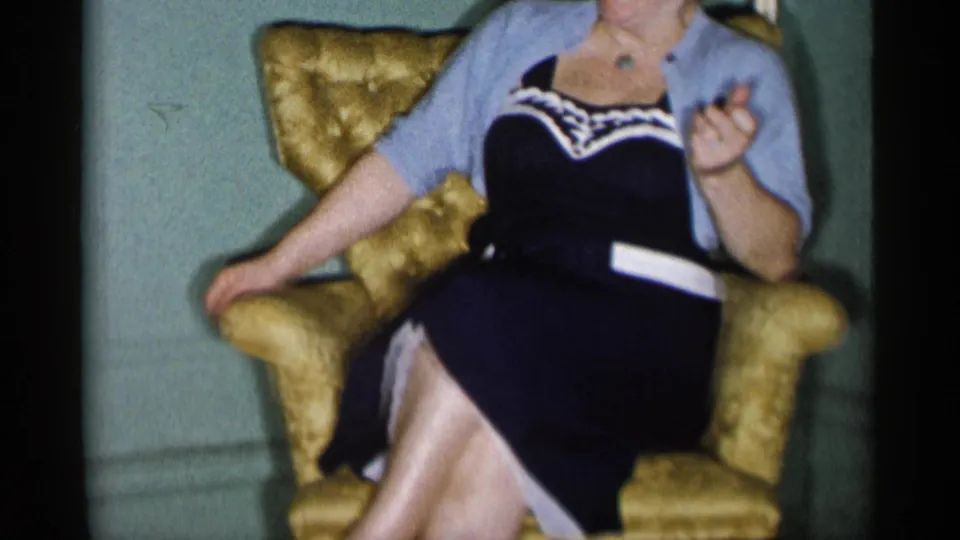 The woman sits in the gold armchair and raises one hand as if speaking or laughing in a 1950s New York City interior.