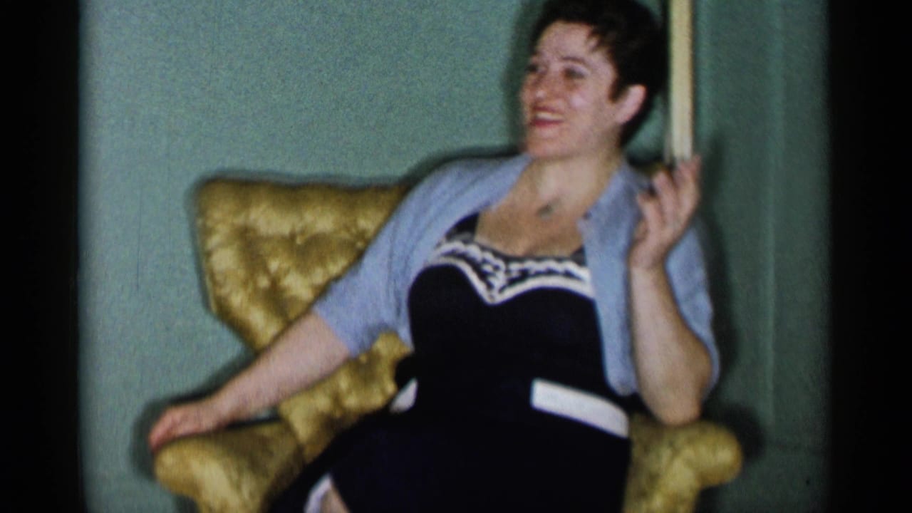 In a 1955 New York City home-movie frame, an adult woman sits smiling in a yellow armchair inside a green-walled room.