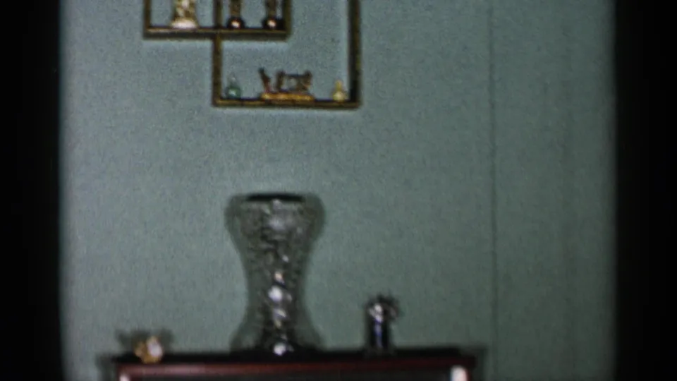 The camera lingers on a 1950s domestic interior with small wall shelves, a glass vase, and a wood-cabinet television set.