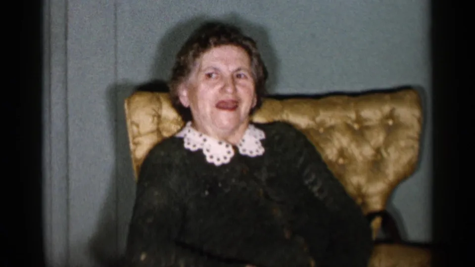 An older adult woman smiles from a yellow armchair in a 1955 New York City home interior, wearing a dark sweater with a white lace collar.