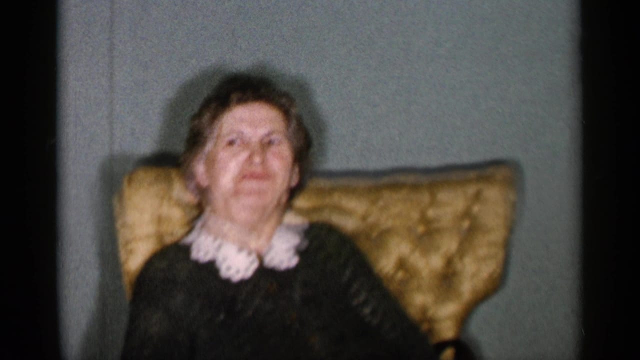 Elderly woman in a dark dress sits in a gold armchair against a pale green wall.