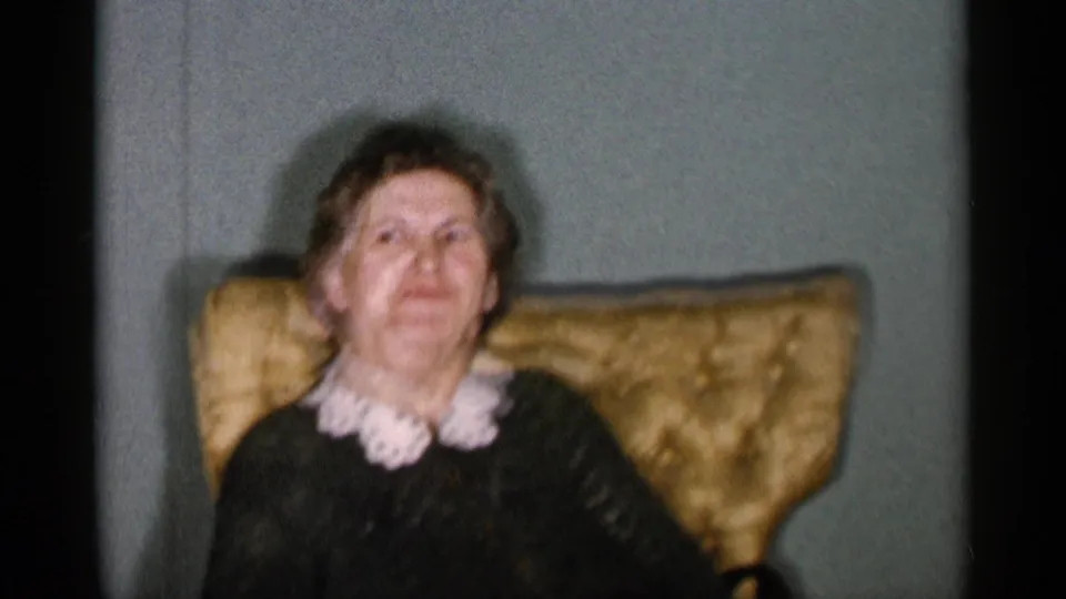 1955 New York City home-movie frame showing an elderly woman seated in a tufted gold armchair, wearing a dark dress with a white lace collar.