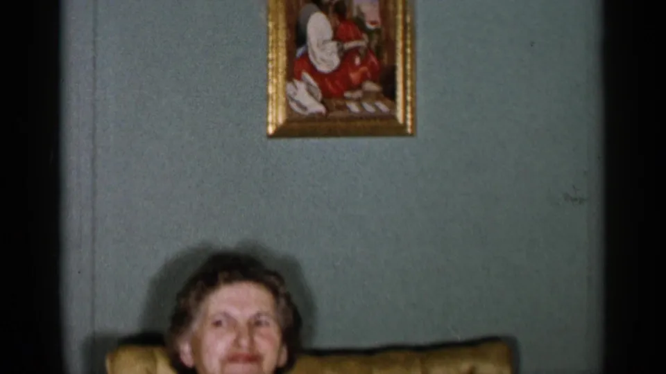 1955 New York City home-movie frame with the seated woman partly lower in view, a gold chair back, pale green wall, and framed painting above.