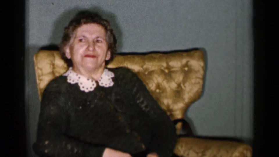 1955 New York City home-movie frame of the elderly woman smiling toward the camera in a dark dress with white collar, seated in a gold armchair.