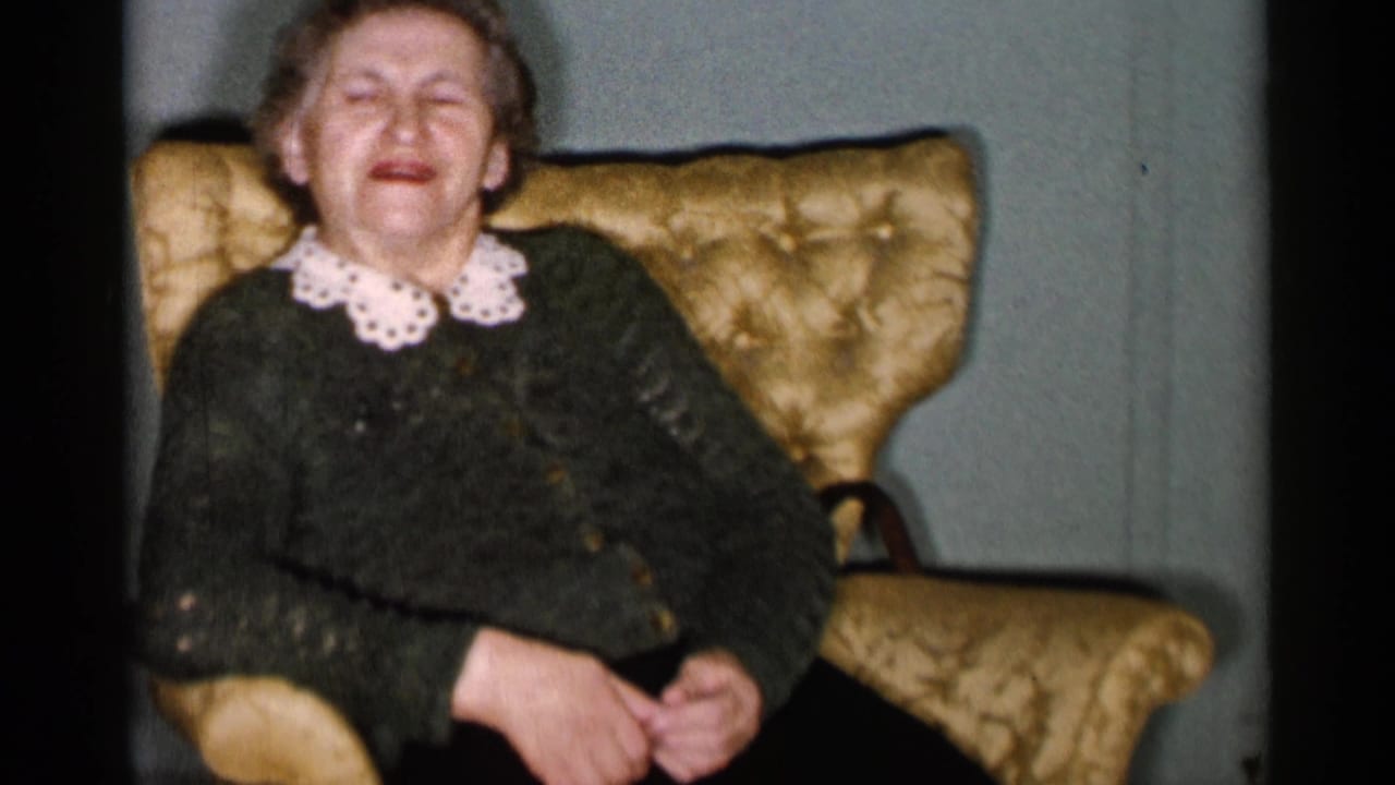 Older woman in a dark cardigan sits smiling in a gold upholstered armchair.