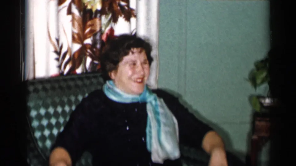 A woman in a black dress and light blue scarf laughs while seated in a patterned chair in a 1955 New York City interior.