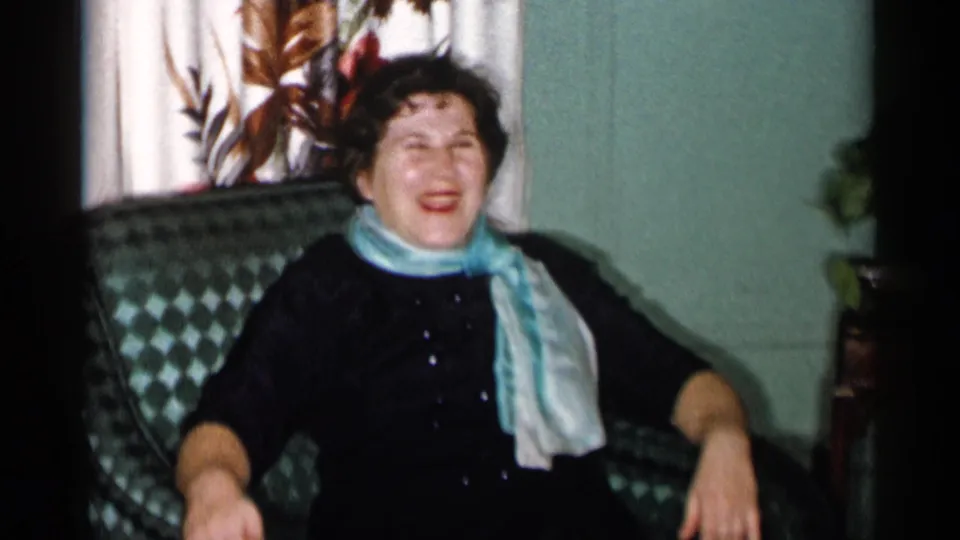 The woman with a light blue scarf laughs in a patterned chair, with curtains and plants visible in the 1955 New York City room.