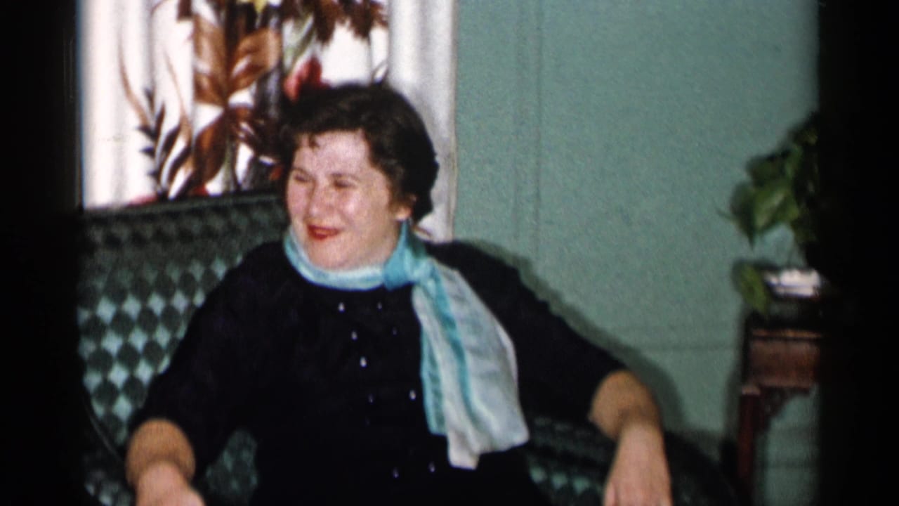 Woman in a dark dress and light scarf seated in a green patterned armchair.