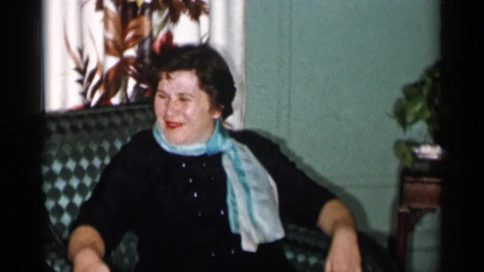 In a 1955 New York City home-movie frame, a woman smiles while seated in a green patterned armchair beside floral curtains.