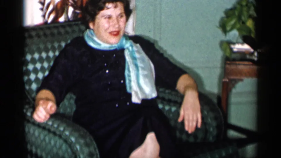 The woman sits back in the patterned green armchair, wearing a dark dress and light scarf in a domestic interior.