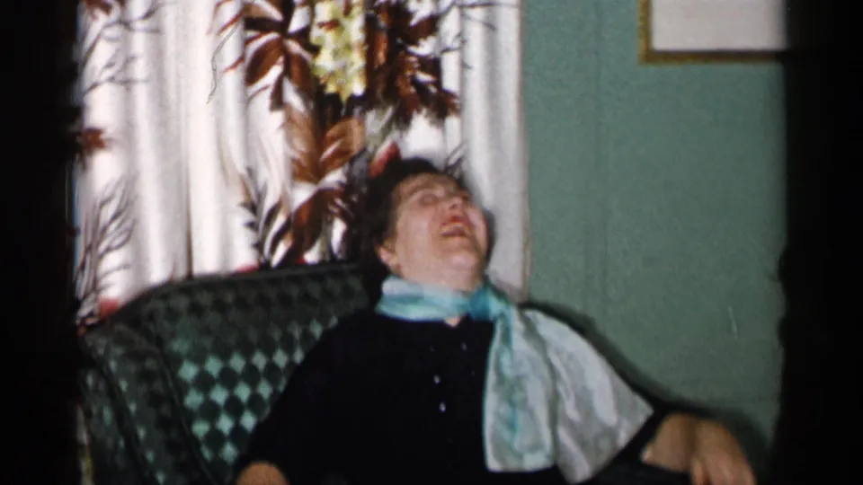 The woman leans back and appears to laugh or speak, with floral curtains and a teal wall behind the armchair.