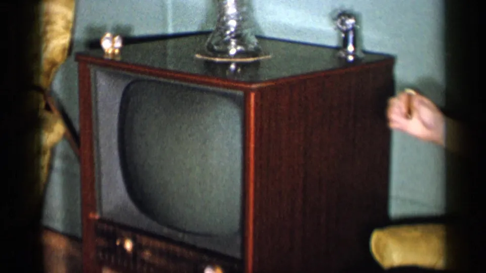 1955 New York City home-movie frame showing a wooden console TV and part of a woman seated nearby in a living room.