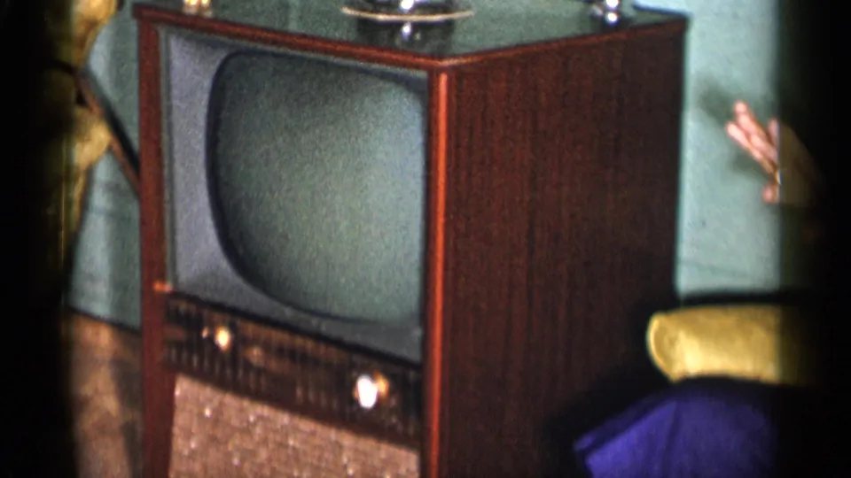 1955 New York City interior frame with a mid-century wooden TV set and a woman partly visible beside an upholstered chair.