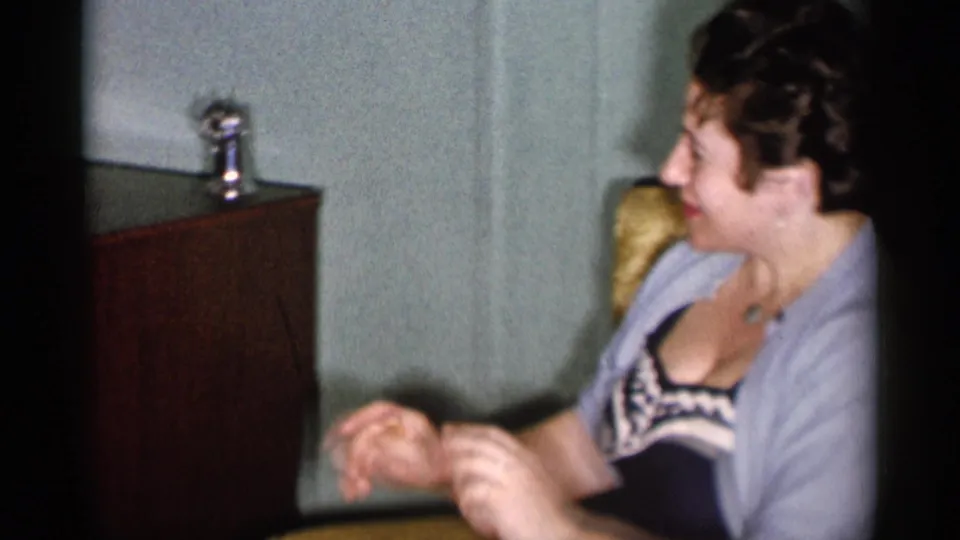 1955 New York City home-movie frame showing a woman in a light cardigan seated beside a wooden TV cabinet.