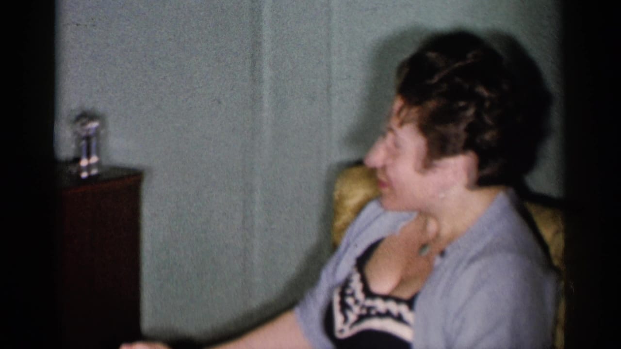 Woman seated beside a dark cabinet with a small object on top in a pastel-walled room.