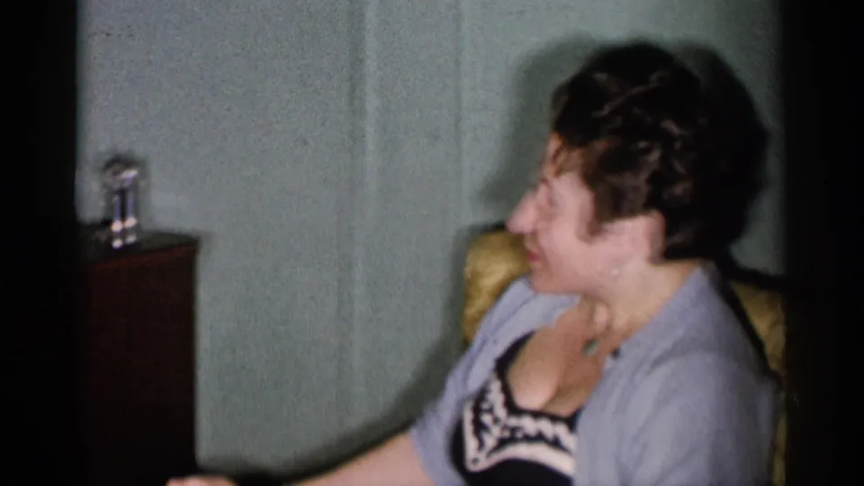 In a 1955 New York City home interior, a seated woman in a cardigan turns beside a dark cabinet and pale wall panels.