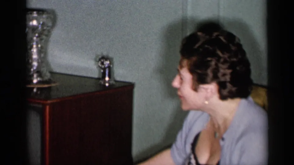 The woman sits in profile in a 1955 New York City interior near a cabinet holding a tall reflective vase and small trophy-like object.