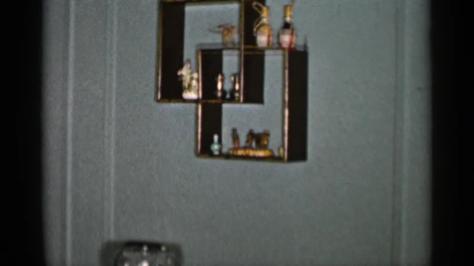 The camera lingers on the geometric display shelf in a 1955 New York City living room, with small ornaments and a vase partly visible below.