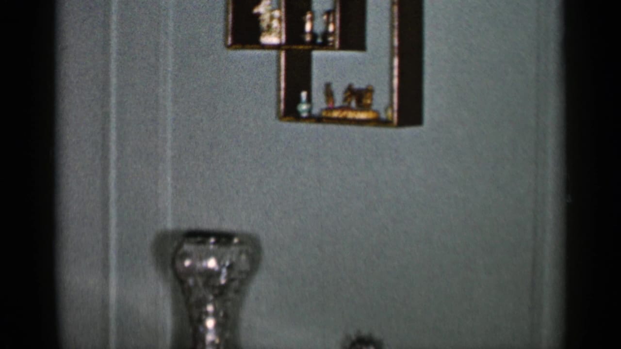 Muted green room wall with shelves, glass vase, chair edge, and dark table.