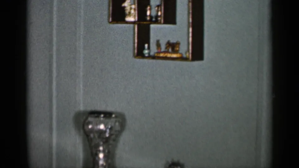 1955 New York City 8mm frame showing a furnished interior with green walls, shelves, a glass vase, and part of a tufted chair.