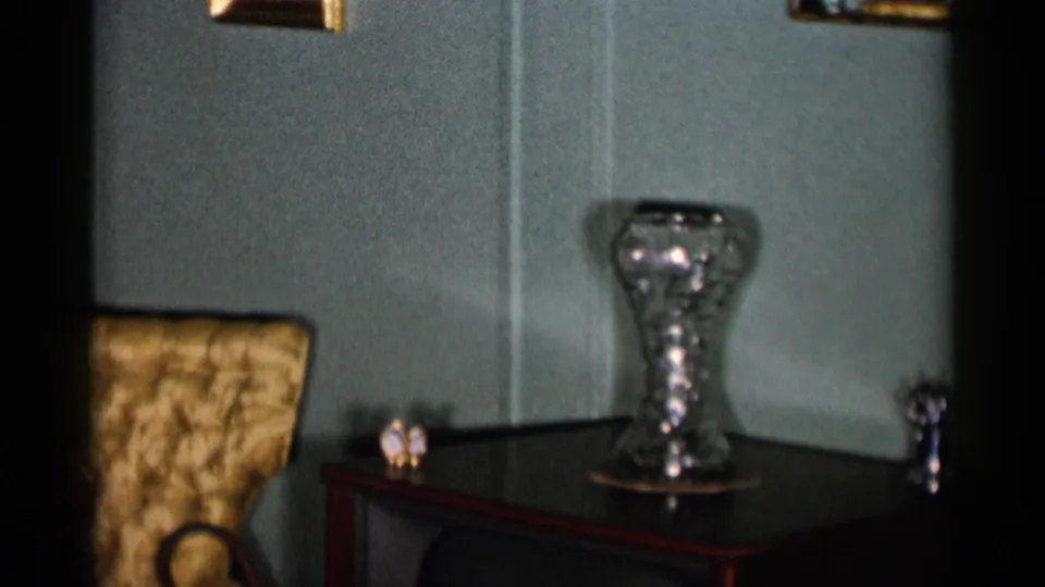 1955 New York City home-movie frame showing a glass vase, dark side table, yellow tufted chair, and muted green interior wall.