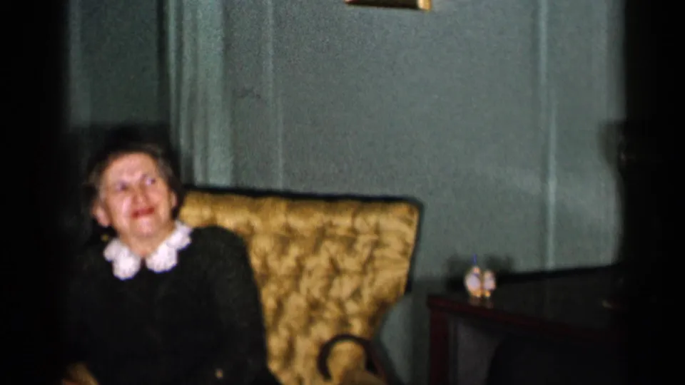 1955 New York City 8mm frame of an adult woman in a dark sweater with white collar seated in a tufted chair indoors.