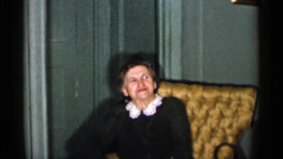 1955 New York City home-movie frame of a seated adult woman looking off camera in a mid-century furnished room.