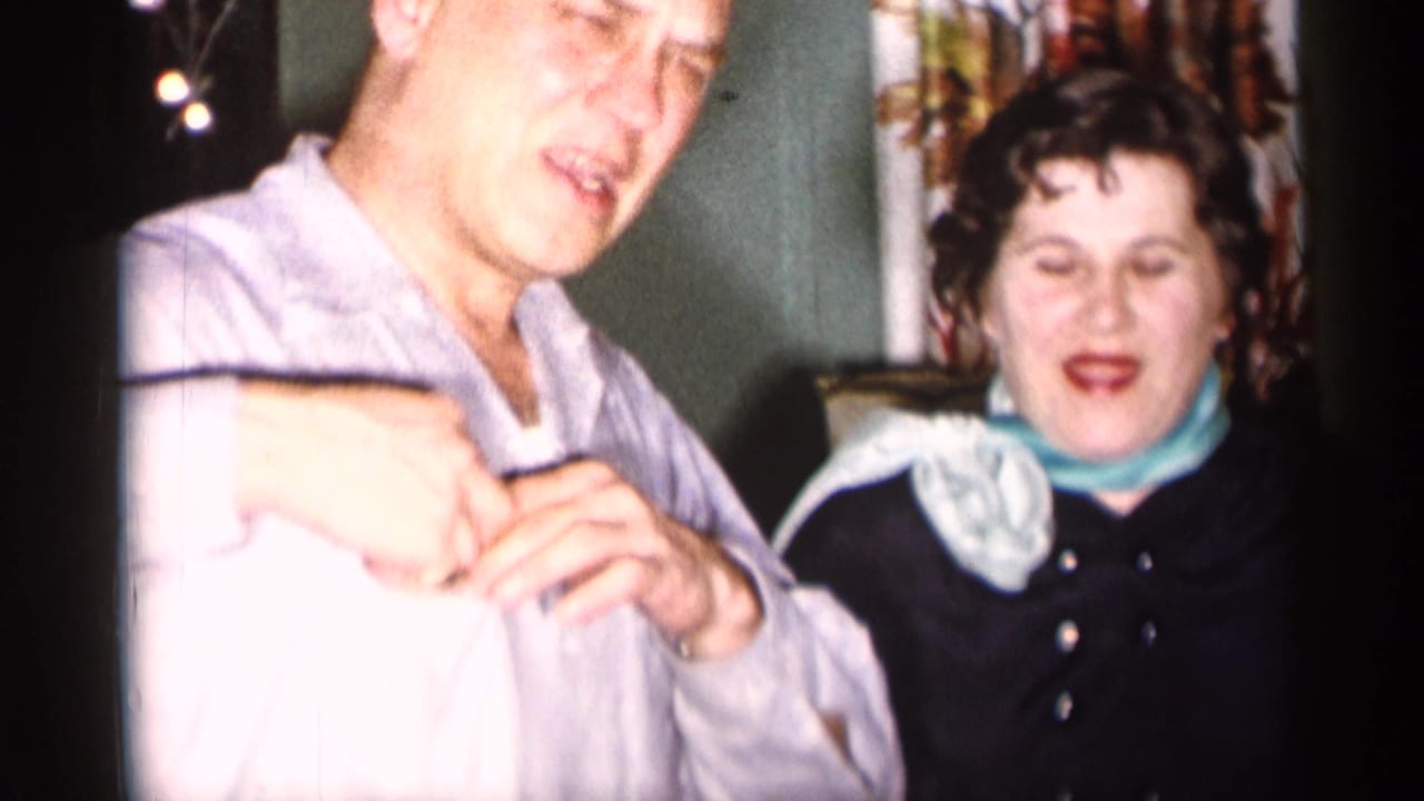 Man in a light shirt stands beside a seated woman in a dark dress and pale scarf.