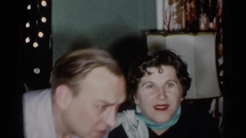 1955 New York home-movie frame with a man bending forward and a seated woman facing the camera in a living room.