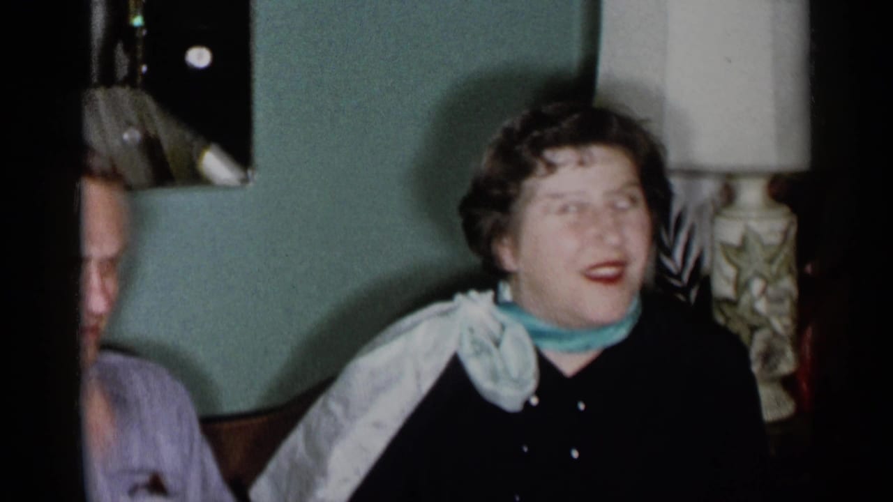 1955 New York City home-movie frame showing a woman on a couch beside a partly cropped man in a dim private living room.