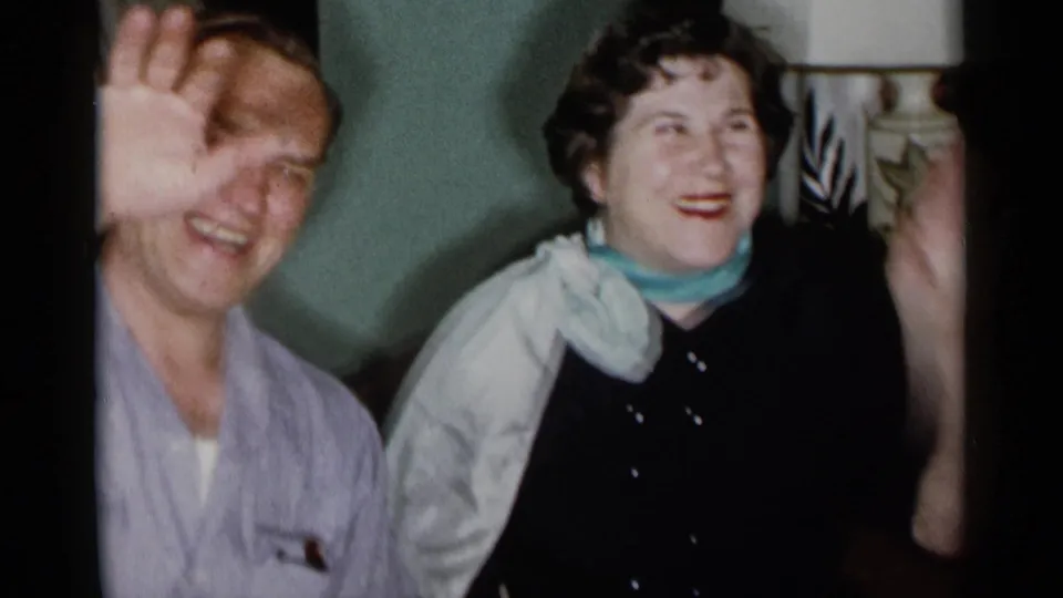 1955 New York City home-movie frame with a man waving and a woman smiling in a private living room.