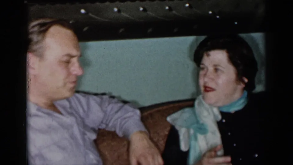 Two adults sit close together in a 1955 New York City living room, with the woman facing the man during conversation.